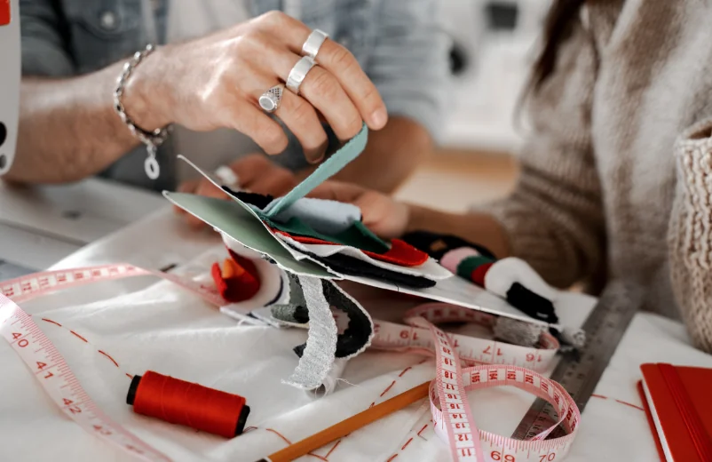Hands trimming fabric swatches with scissors beside thread spools and a measuring tape