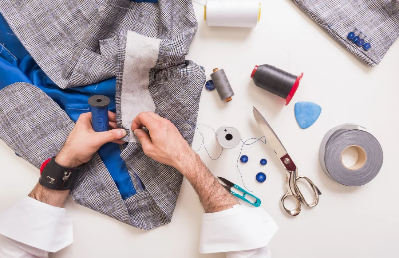 Tailor working on a grey jacket surrounded by thread, scissors and buttons