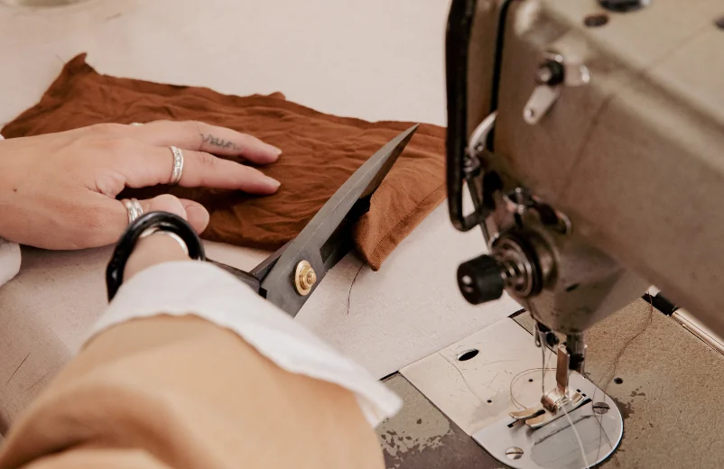 Hands cutting brown fabric with tailor's shears next to a sewing machine