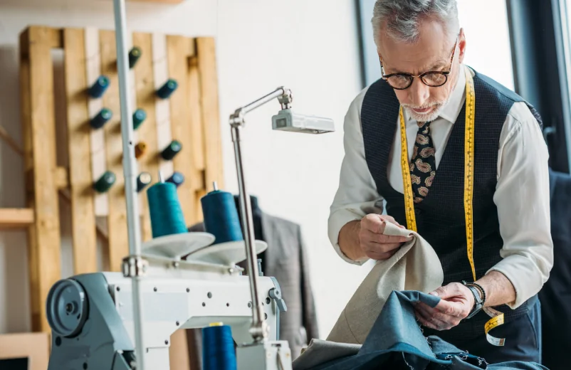 Tailor examining a blue garment at his workbench with a tape measure around his neck