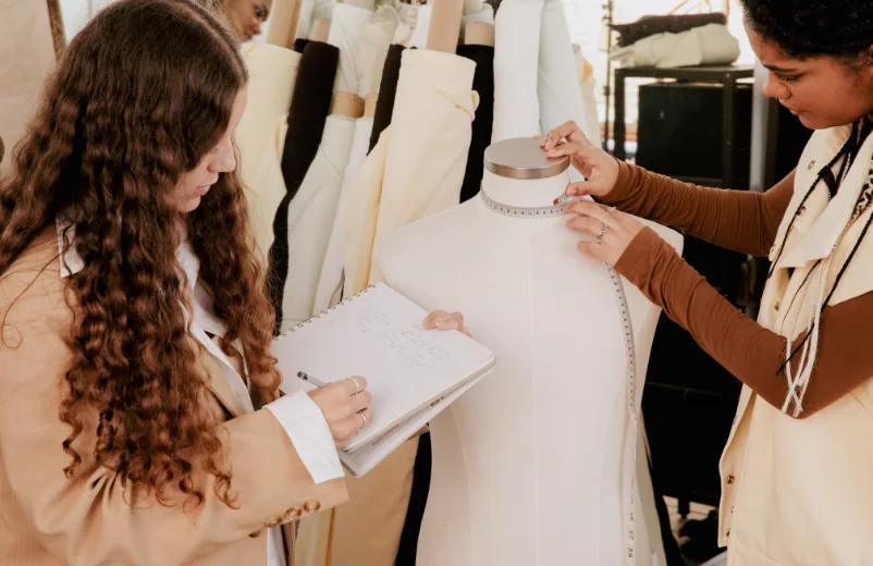 Two designers draping fabric and taking notes on a dress form